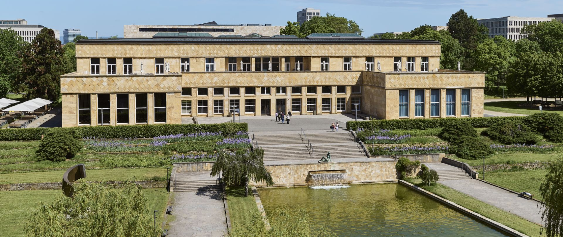 Historisches Campusgebäude mit heller Sandsteinfassade, großen Fenstern und breiter Freitreppe, davor ein Wasserbecken mit Fontäne und gepflegten Gartenanlagen; links eine Außenterrasse mit weißen Sonnenschirmen – idealer Rahmen für Veranstaltungen auf dem Campus Westend.