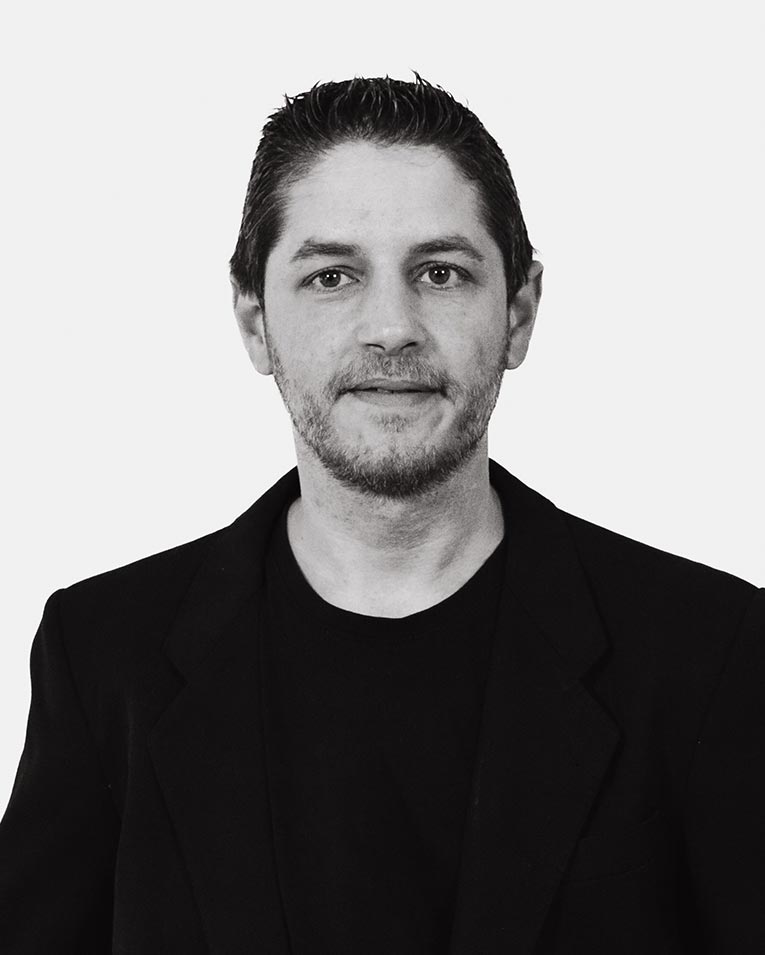 Schwarz-weiß Porträt eines Mannes mit kurzem dunklem Haar und leichtem Bartschatten, der ein dunkles Sakko und T-Shirt trägt, Teammitglied des Catering-Unternehmens