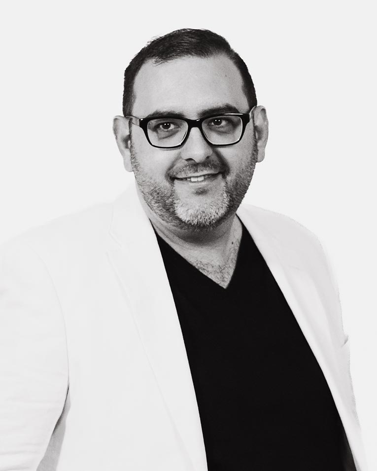 Schwarz-weiß Porträt eines lächelnden Mannes mit Brille und Bart, in weißem Blazer und dunklem Hemd, Teammitglied des Catering-Unternehmens