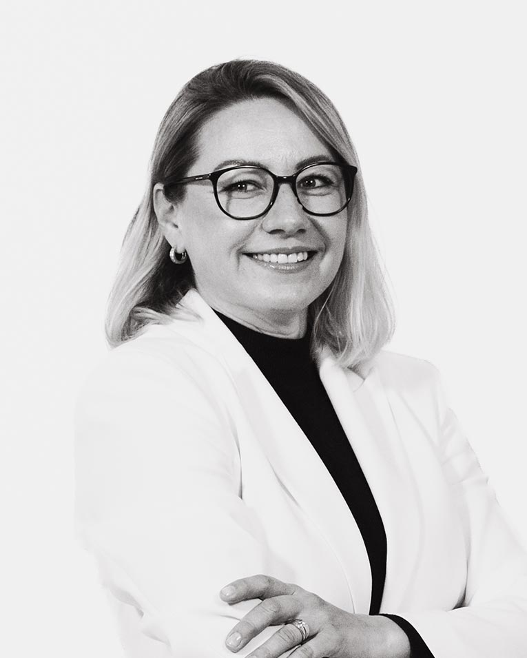 Schwarz-weiß Porträt einer lächelnden Frau mit schulterlangen blonden Haaren und Brille, in weißem Blazer mit verschränkten Armen, Teammitglied des Catering-Unternehmens