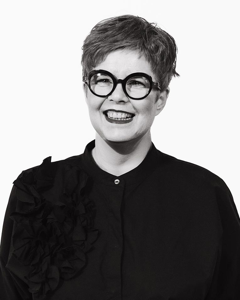 Schwarz-weiß Porträt einer fröhlich lächelnden Frau mit kurzen Haaren, runden Brillen und schwarzer Bluse mit Rüschen, Teammitglied des Catering-Unternehmens