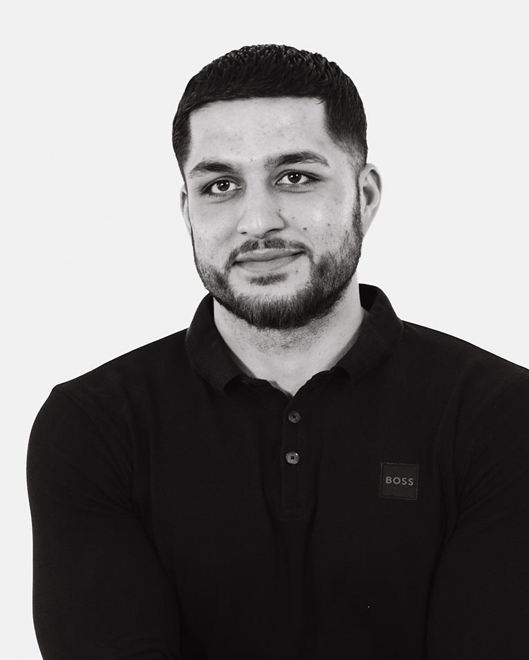 Schwarz-weiß Porträt eines jungen Mannes mit kurzem Haar und Bart, der ein dunkles Polohemd mit Logo trägt und lächelt, Teammitglied des Catering-Unternehmens